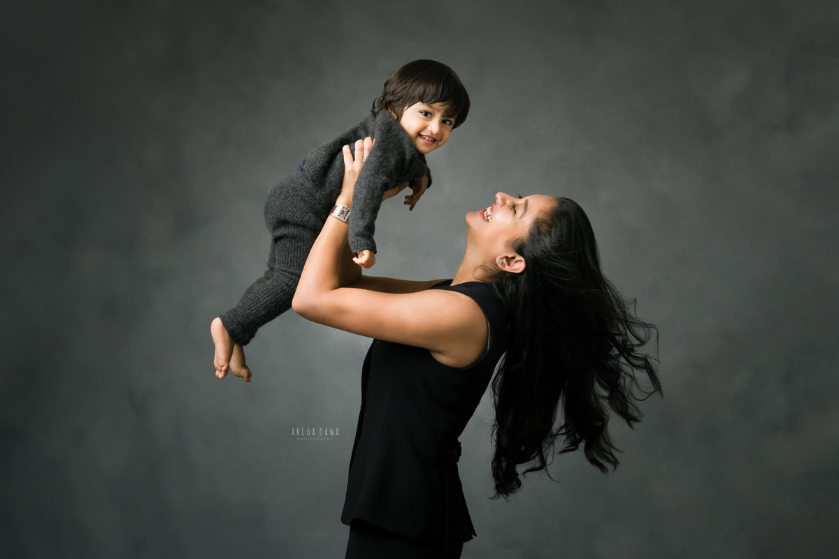 1-year boy lifted in mom's arms against a serene grey backdrop, beautifully captured by Anega Bawa Photography, the premier choice for family photo shoots in Gurgaon (Delhi NCR).