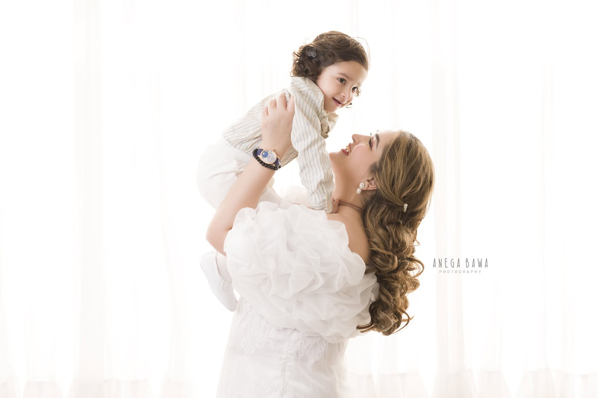 1-year-old boy lifted in mom's arms with a white backdrop, capturing a loving moment. This family photo shoot is done by Anega Bawa Photography in Gurgaon (Delhi NCR), ensuring every smile is cherished.