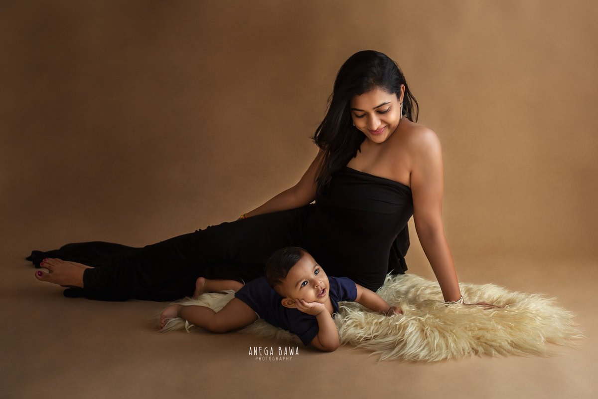 Cute 1-year-old boy lying on a beige rug with mom against a brown backdrop, captured by Anega Bawa Family Photographer Gurgaon (Delhi NCR).