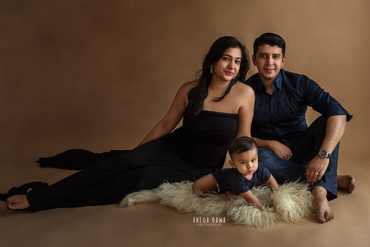 1-year-old boy enjoying a serene family photo shoot on a beige rug with his loving parents, captured by Anega Bawa Photography in Gurgaon (Delhi NCR). The warm brown backdrop adds to the intimate and stylish atmosphere of the session.