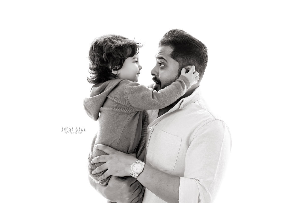 Adorable 1-year-old boy playing with dad's ears and posing cutely against a white backdrop, captured by Anega Bawa Family Photographer Gurgaon (Delhi NCR).