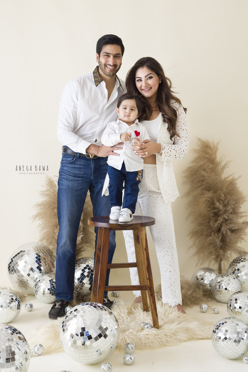 1-year-old boy poses cutely with mom and dad in front of bushes and disco balls on a beige backdrop, family photoshoot in Gurgaon (Delhi NCR)