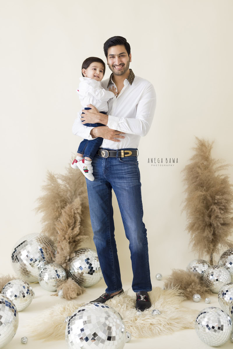 1-year-old boy poses with dad amidst bushes and disco balls against a beige backdrop, family photoshoot in Gurgaon (Delhi NCR)