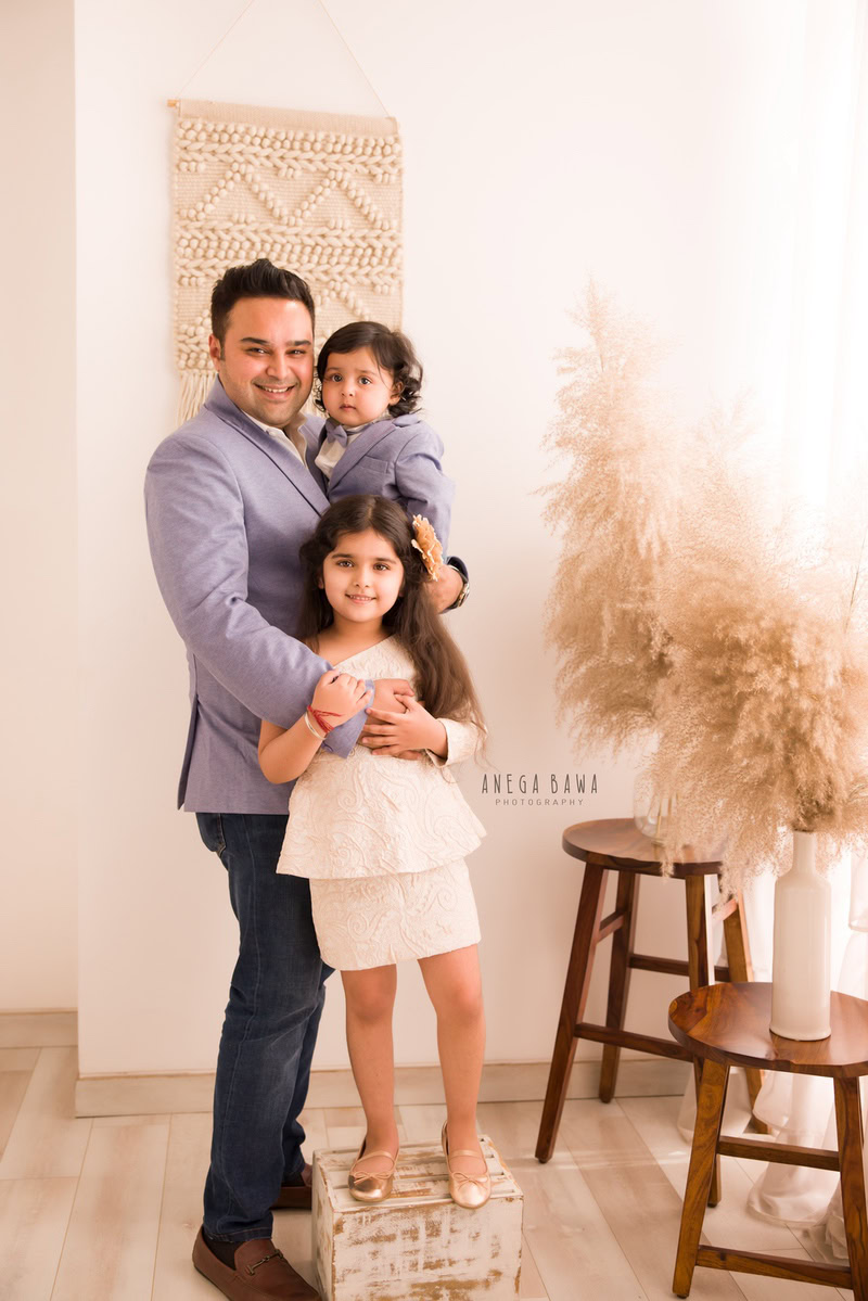 1-year-old boy posing with dad and elder sister against a backdrop of bushes and beige background, beautifully captured by Anega Bawa Photography during a family photoshoot in Gurgaon (Delhi NCR).