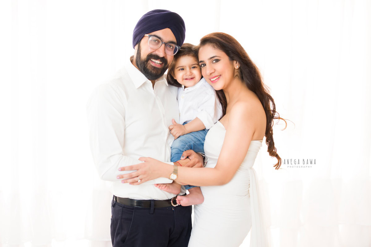 1-year-old boy poses with mom and dad, all wearing white, against a serene white backdrop, captured by Anega Bawa Photography for a family photoshoot in Gurgaon (Delhi NCR).