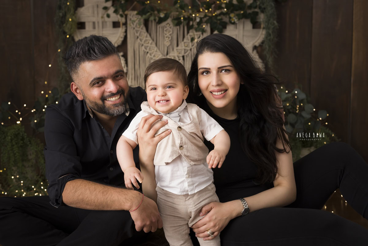 1-year-old boy poses with mom and dad against a warm brown backdrop adorned with fairy lights, beautifully captured by Anega Bawa Photography for a family photoshoot in Gurgaon (Delhi NCR).