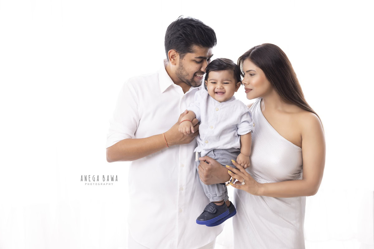 1-year-old boy posing in a cute, smiling pose with mom and dad against a white backdrop. This family photo shoot is done by Anega Bawa Photography in Gurgaon (Delhi NCR) to capture these joyful moments.