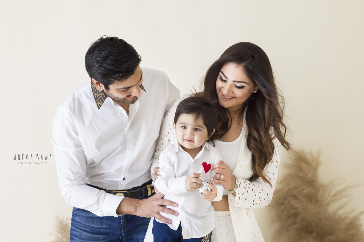 1-year-old boy posing cutely with mom and dad against a beige backdrop with bushes, captured by Anega Bawa Photography for a family photoshoot in Gurgaon (Delhi NCR).
