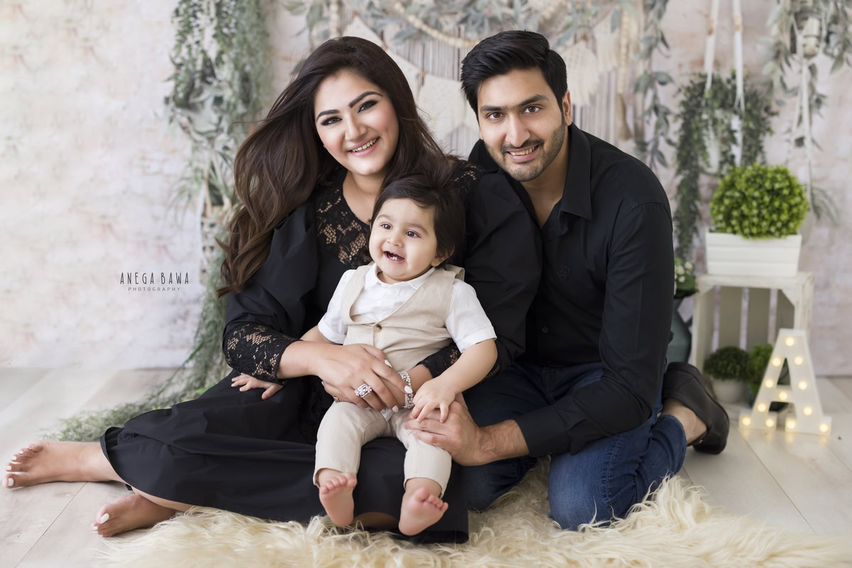 1-year-old boy posing cute with mom and dad wearing black, surrounded by leafy fringes against a beige backdrop. This beautiful family photo shoot is done by Anega Bawa Photography in Gurgaon (Delhi NCR), capturing cherished moments.