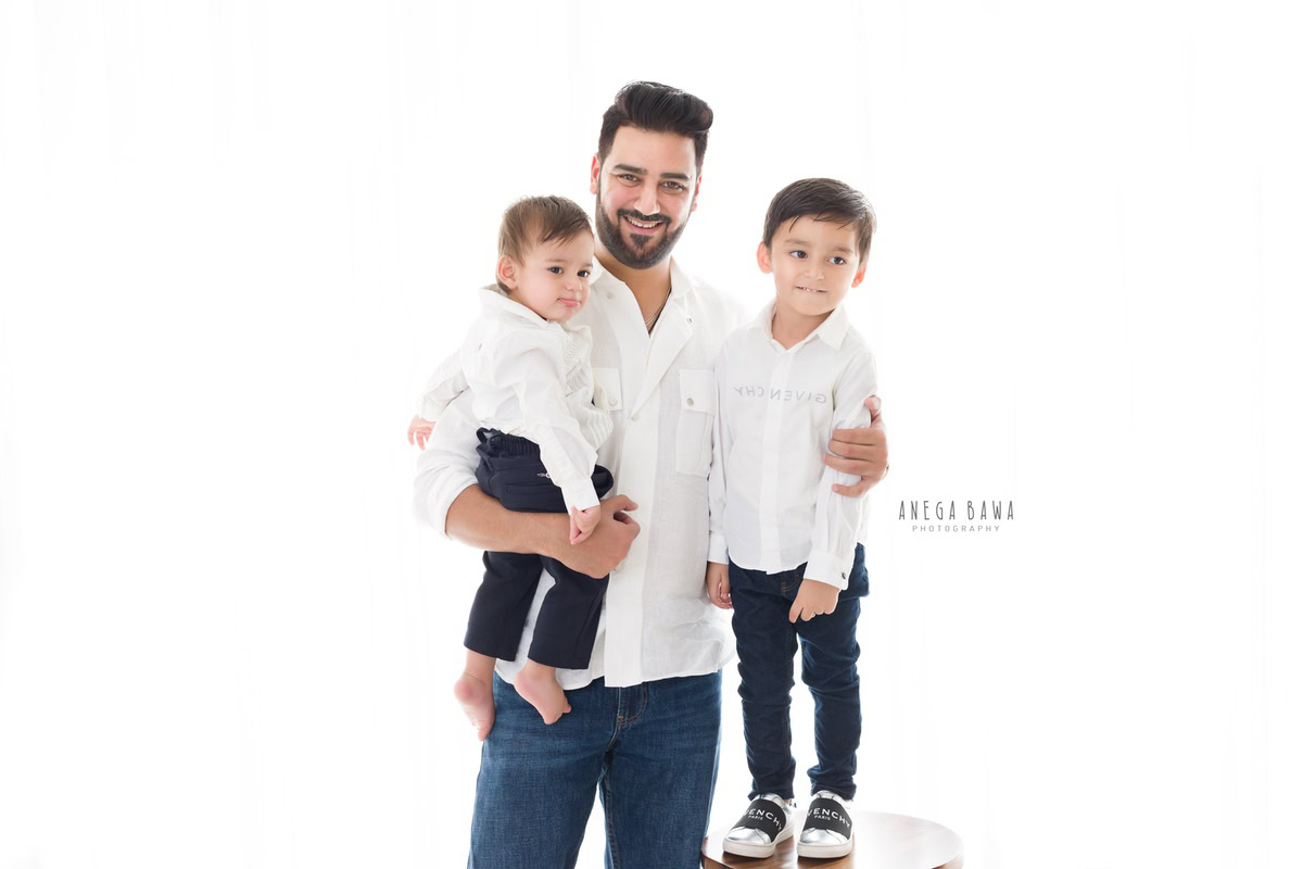 1-year-old boy posing with dad and elder son, all dressed in white shirts and denims, against a clean white backdrop. Captured by Anega Bawa Photography, Gurgaon (Delhi NCR).