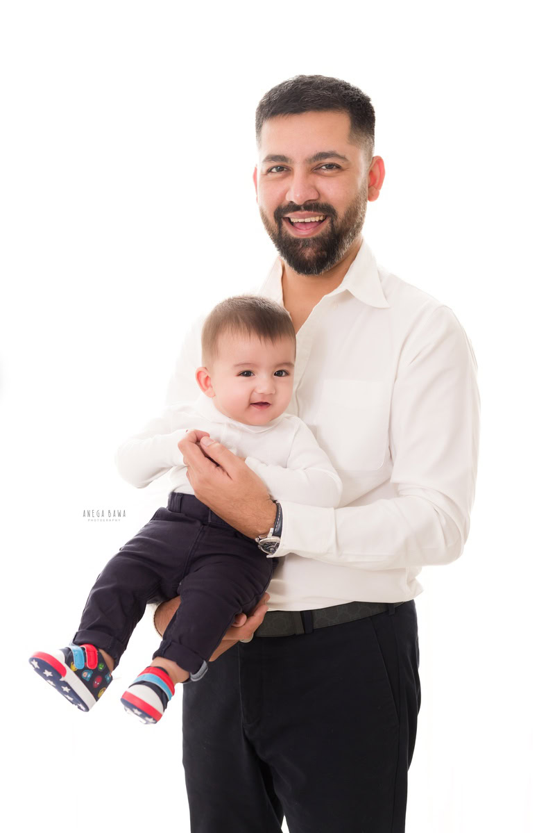 1-year-old boy posing with dad, both twinning in white shirts and black trousers, against a clean white backdrop. Captured by Anega Bawa Photography, Gurgaon (Delhi NCR).
