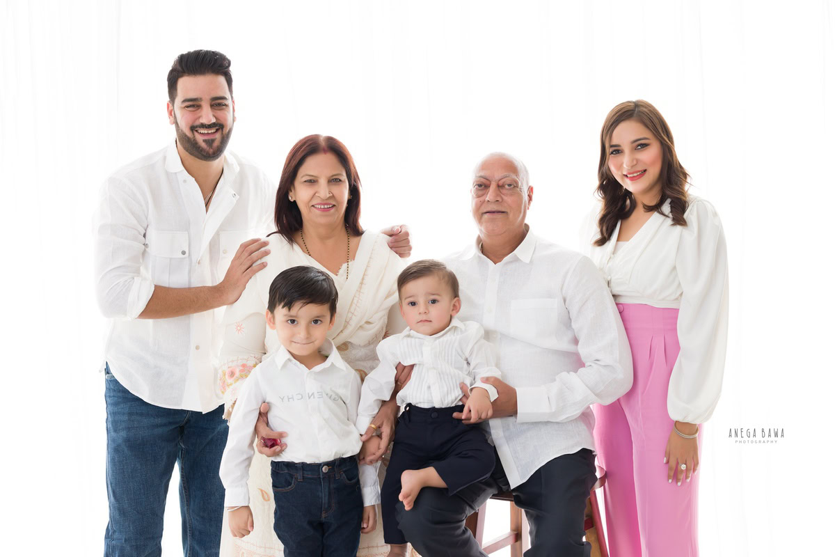 1-year-old boy posing with grandparents, mom, dad, and elder brother in a family pose against a white backdrop, all wearing white shirts. Captured by Anega Bawa Photography, Gurgaon (Delhi NCR).