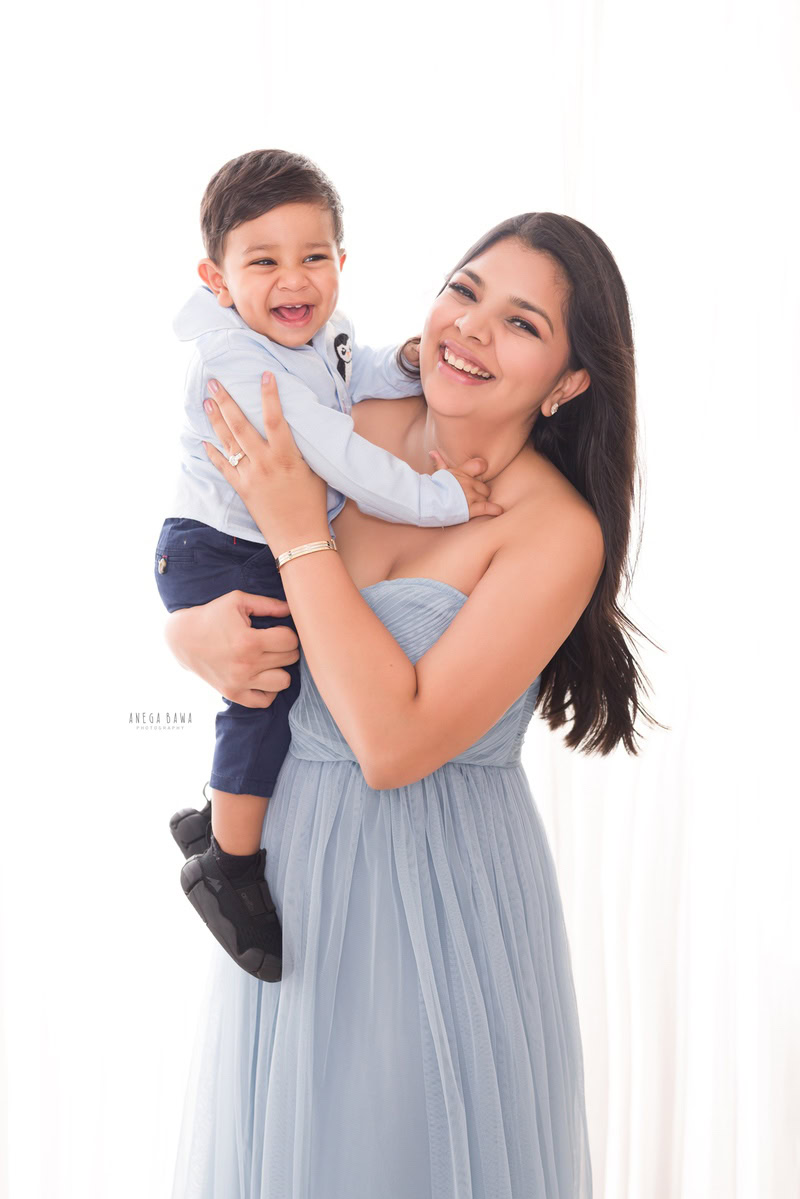 1-year-old boy posing with mom in a cute smiling pose against a white backdrop. Captured by Anega Bawa Photography, Gurgaon (Delhi NCR).