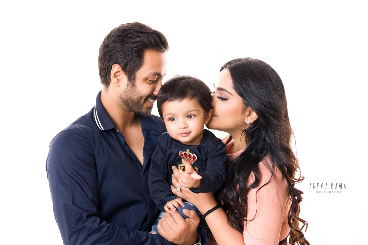 1-year-old boy posing with mom and dad in a cute kissing pose against a white backdrop. Captured by Anega Bawa Photography, Gurgaon (Delhi NCR).
