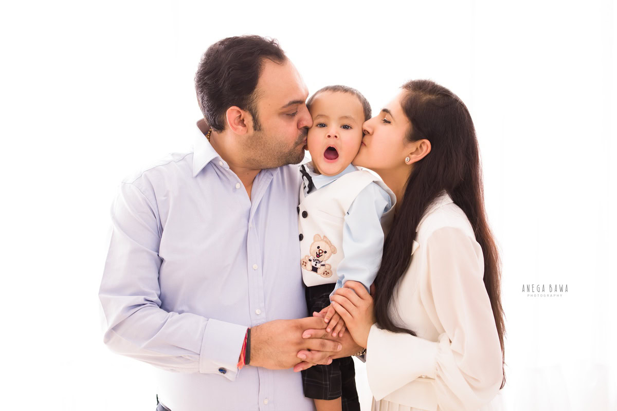 1-year-old boy posing with mom and dad in a kissing pose against a white backdrop. Captured by Anega Bawa Photography, Gurgaon (Delhi NCR).