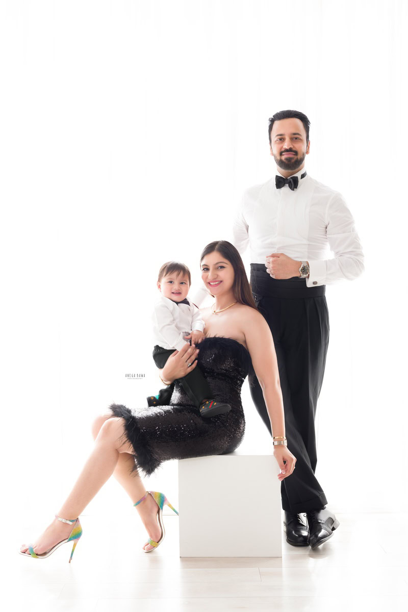 1-year-old boy posing with mom and dad against a white backdrop. Son and dad wearing white shirt and black bow, captured by Anega Bawa, family photographer in Gurgaon (Delhi NCR).