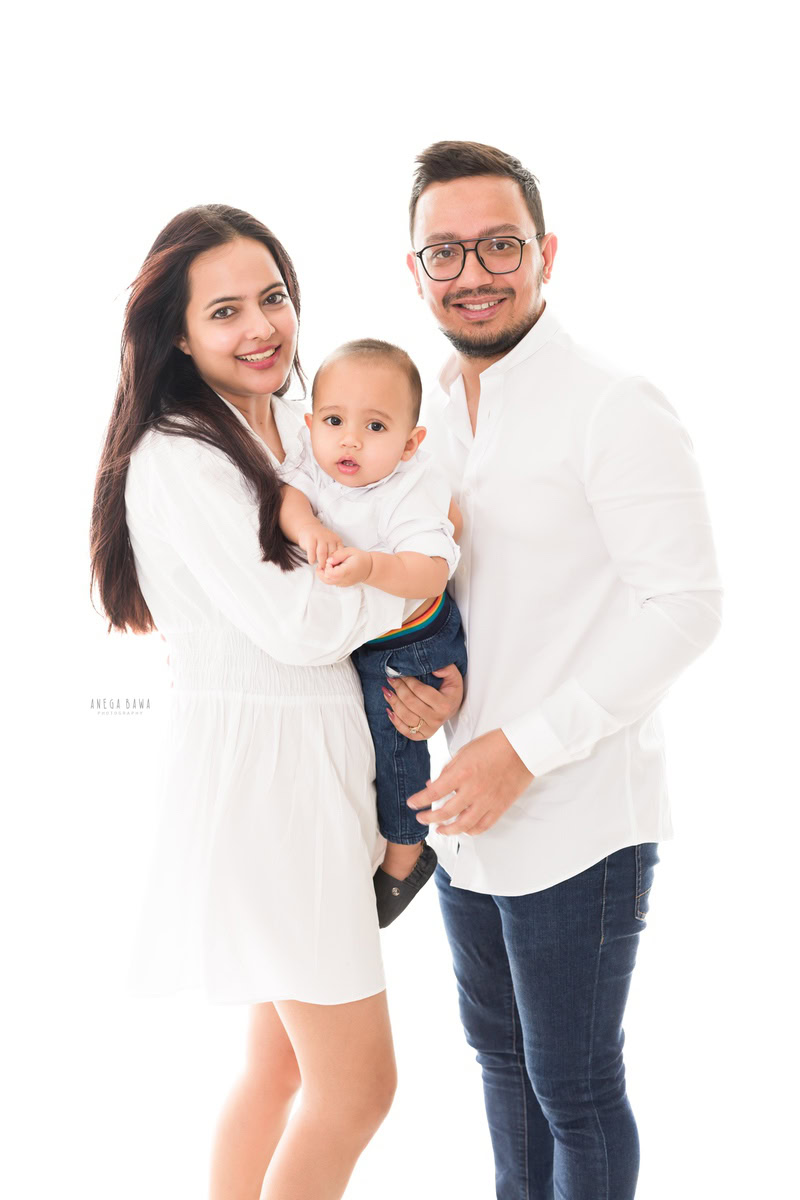 1-year-boy posing with mom and dad against a clean white backdrop, all dressed in elegant white attire, captured by Anega Bawa Photography for a family photoshoot in Gurgaon (Delhi NCR).