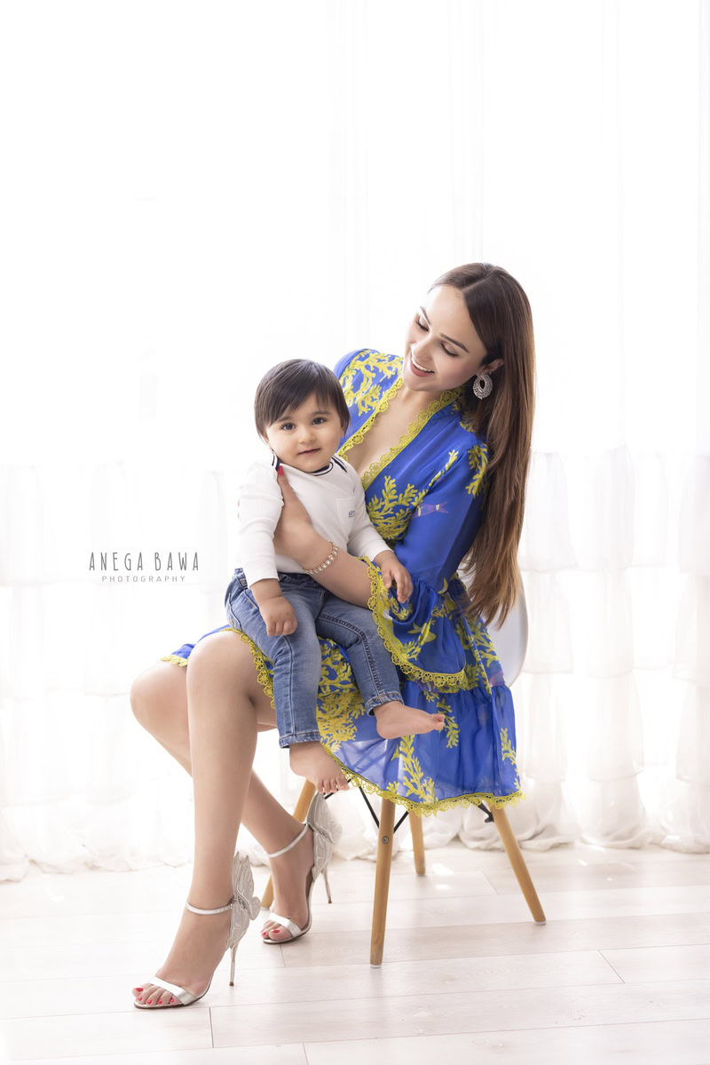 Impressive 1-year-old boy posing with mom seated on a stylish stool against a pristine white backdrop, captured by Anega Bawa Family Photographer Gurgaon (Delhi NCR).