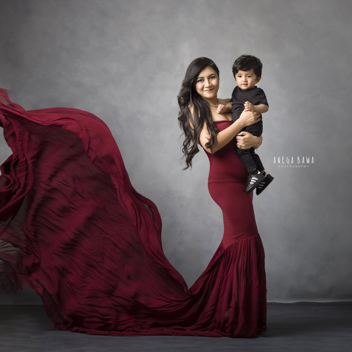 1-year-old boy posing with mom, who is wearing a red gown, against a grey backdrop, family photoshoot in Gurgaon (Delhi NCR)