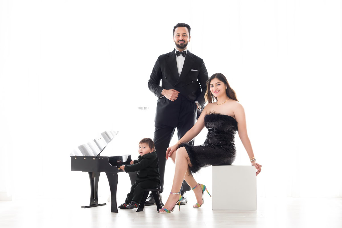 1-year-old boy posing with a piano, dressed in a black suit, with mom and dad, all wearing black, against a white backdrop, captured by Anega Bawa Family Photographer Gurgaon (Delhi NCR).