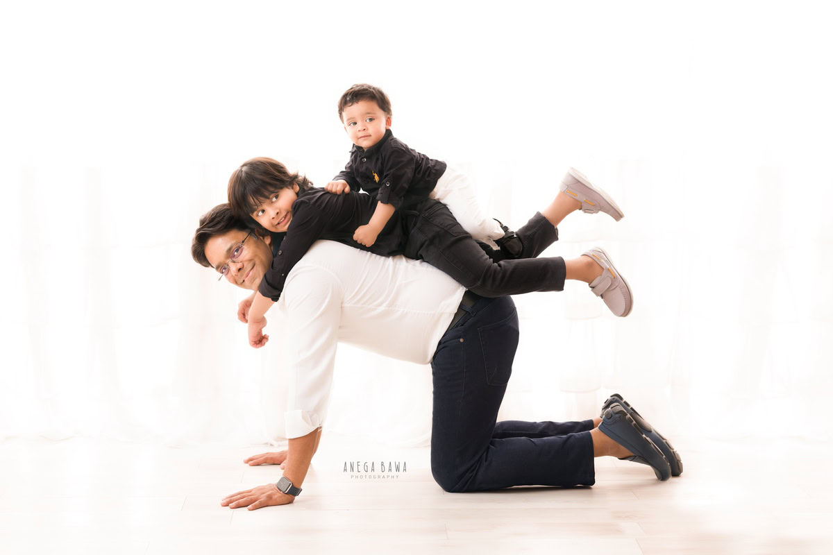 1-year-old boy sitting on dad's back, with elder brother posing, against a white backdrop, family photoshoot in Gurgaon (Delhi NCR)