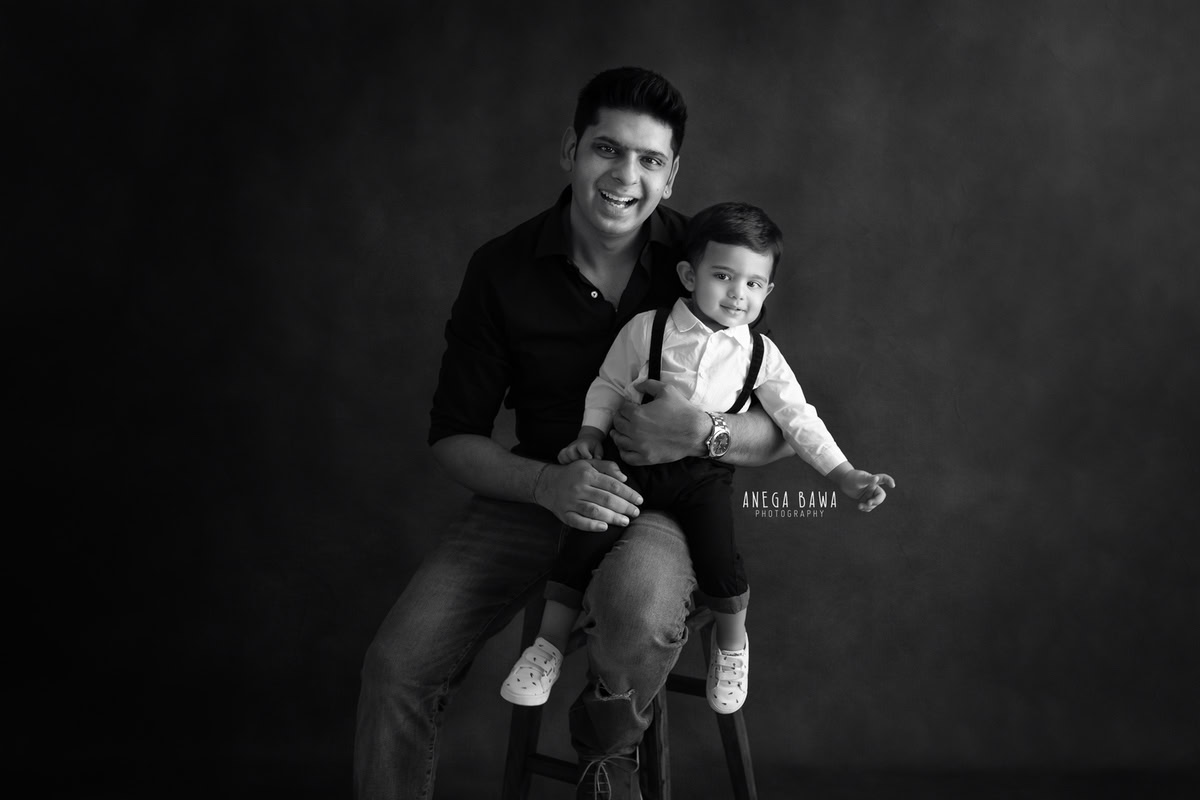 1-year-old boy seated on dad's lap against a black backdrop, captured by Anega Bawa Photography for a family photoshoot in Gurgaon (Delhi NCR).