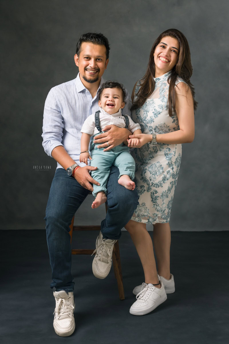 1-year-old boy sitting on dad's lap in a cute smiling pose, with mom standing, against a grey backdrop, family photoshoot in Gurgaon (Delhi NCR)