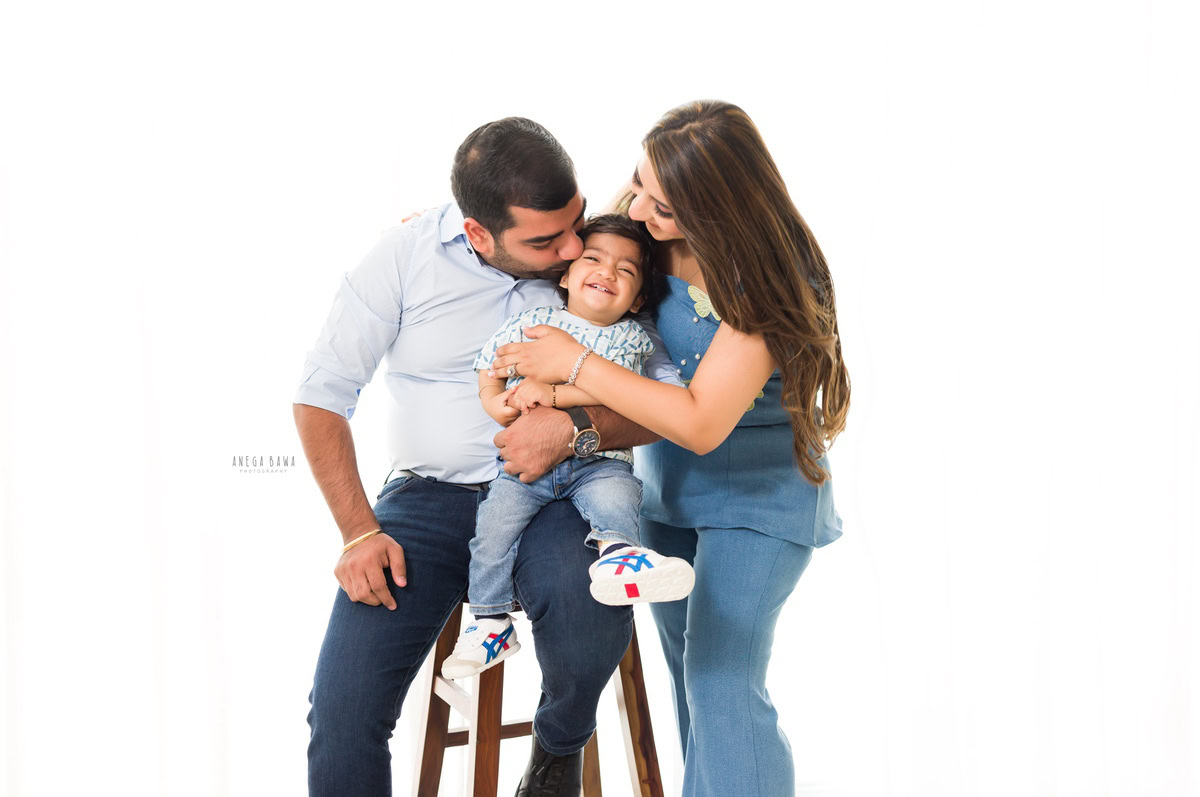 1-year-boy seated on dad's lap with mom kissing him against a white backdrop, captured by Anega Bawa Photography for a family photoshoot in Gurgaon (Delhi NCR).