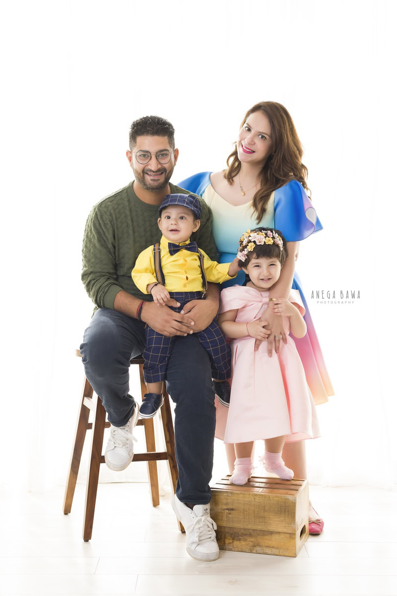1-year-old boy seated on dad's lap, mom posing with elder daughter against a white backdrop, captured by Anega Bawa Family Photographer Gurgaon (Delhi NCR).
