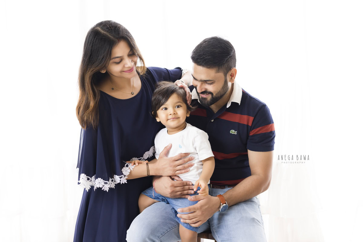 1-year-old boy seated on dad's lap with mom posing on a white backdrop, captured by Anega Bawa Family Photographer in Gurgaon (Delhi NCR).