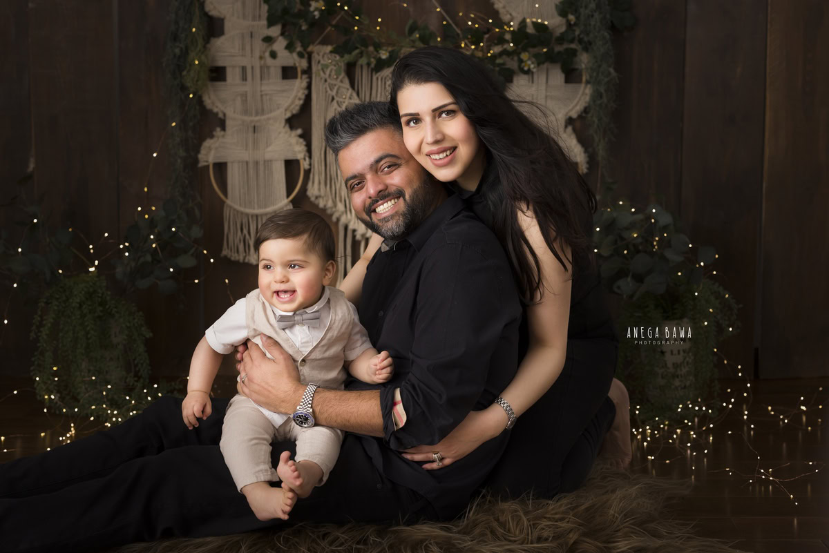 1-year-old boy seated on dad's lap, posing cutely with mom hugging from behind, against a backdrop of fairy lights and brown backdrop, captured by Anega Bawa, family photographer in Gurgaon (Delhi NCR).