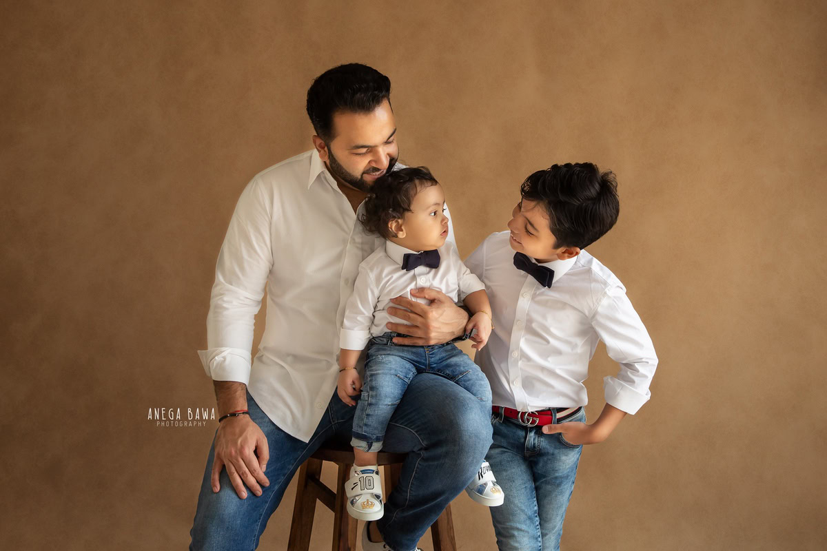 1-year-old boy seating on dad's lap, posing with elder brother against a brown backdrop, captured by Anega Bawa, family photographer in Gurgaon (Delhi NCR).