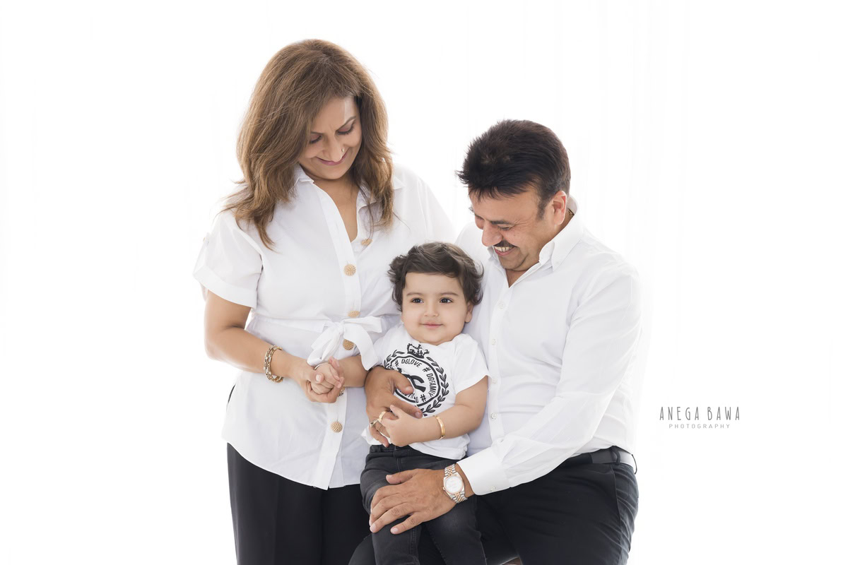 1-year-boy seated on dad's lap, posing with mom, all wearing white against a serene white backdrop, beautifully captured by Anega Bawa Photography for a family photoshoot in Gurgaon (Delhi NCR).