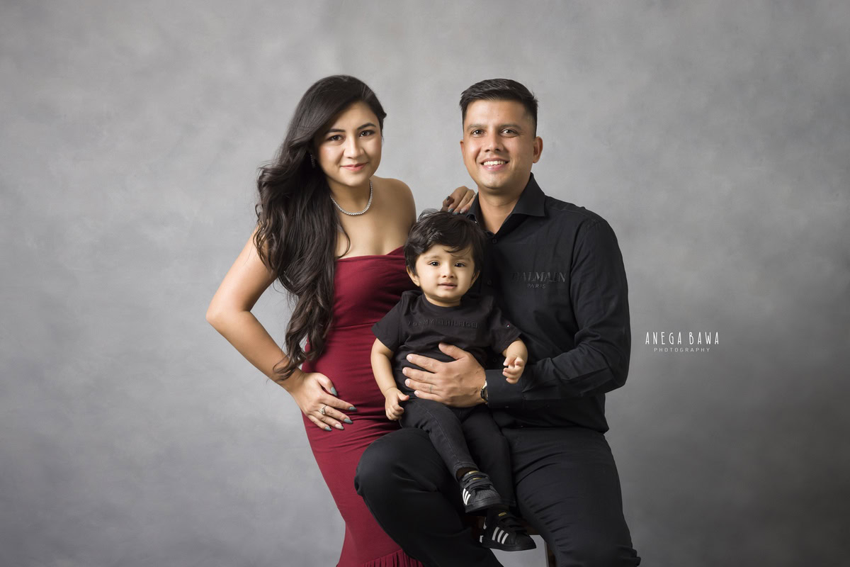 1-year boy sitting on dad's lap, twinning in black shirts, posing with mom against a grey backdrop, captured by Anega Bawa Photography for a family photoshoot in Gurgaon (Delhi NCR).