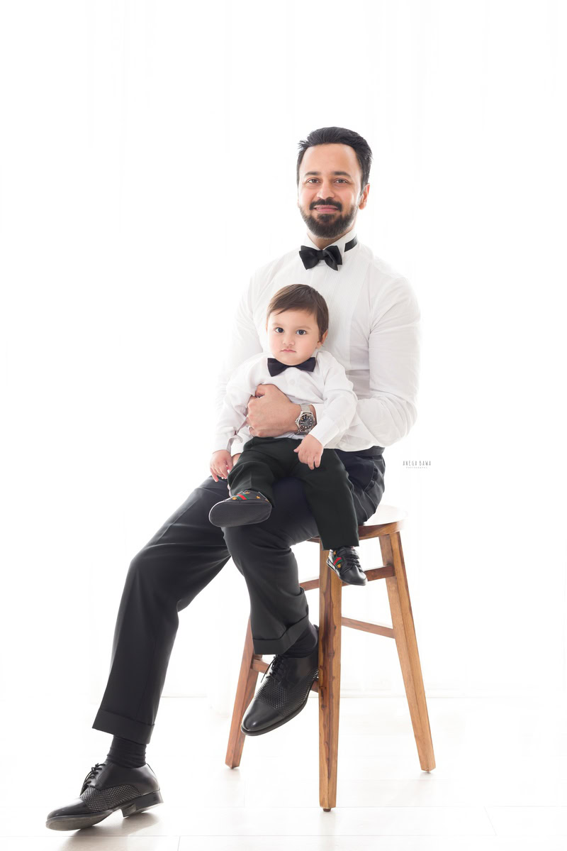 1-year-old boy seating on dad's lap, twinning in white shirt and black trousers with a black bow tie against a white backdrop, captured by Anega Bawa, family photographer in Gurgaon (Delhi NCR).