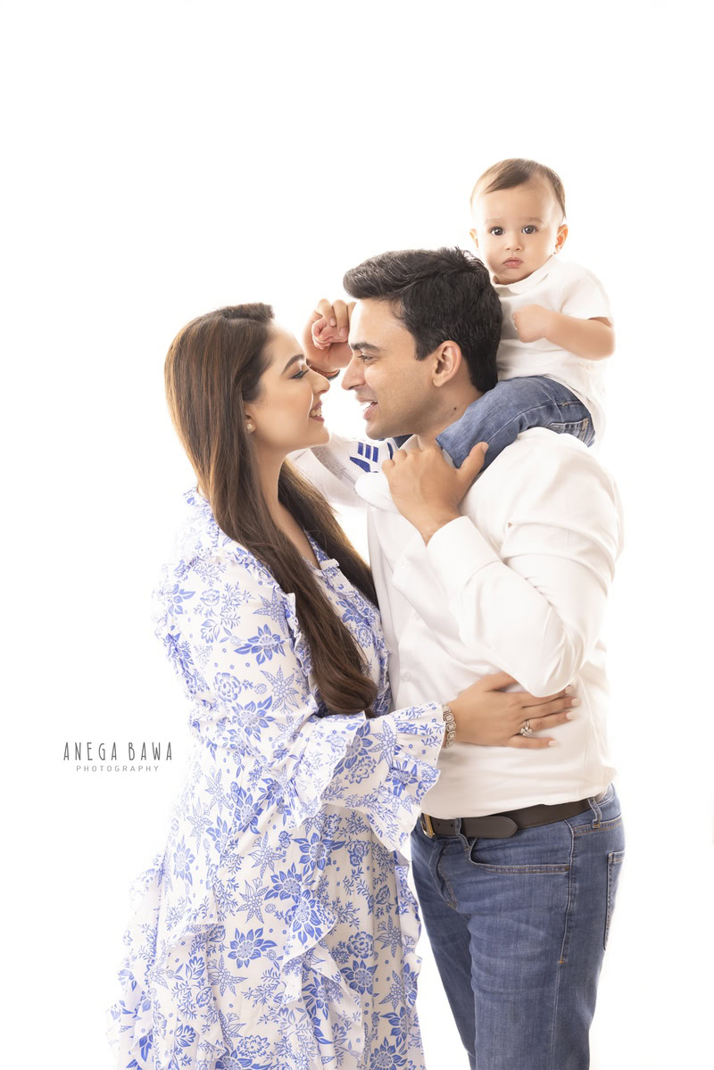 1-year-boy seated on dad's shoulder, posing with mom against a clean white backdrop, captured by Anega Bawa Photography for a family photoshoot in Gurgaon (Delhi NCR).
