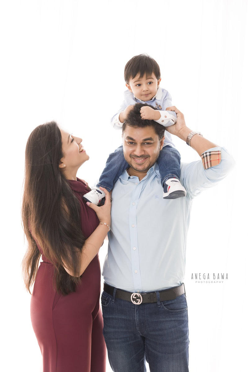 1-year-old boy seated on dad's shoulders, posing with mom against a white backdrop, captured by Anega Bawa Family Photographer Gurgaon (Delhi NCR).