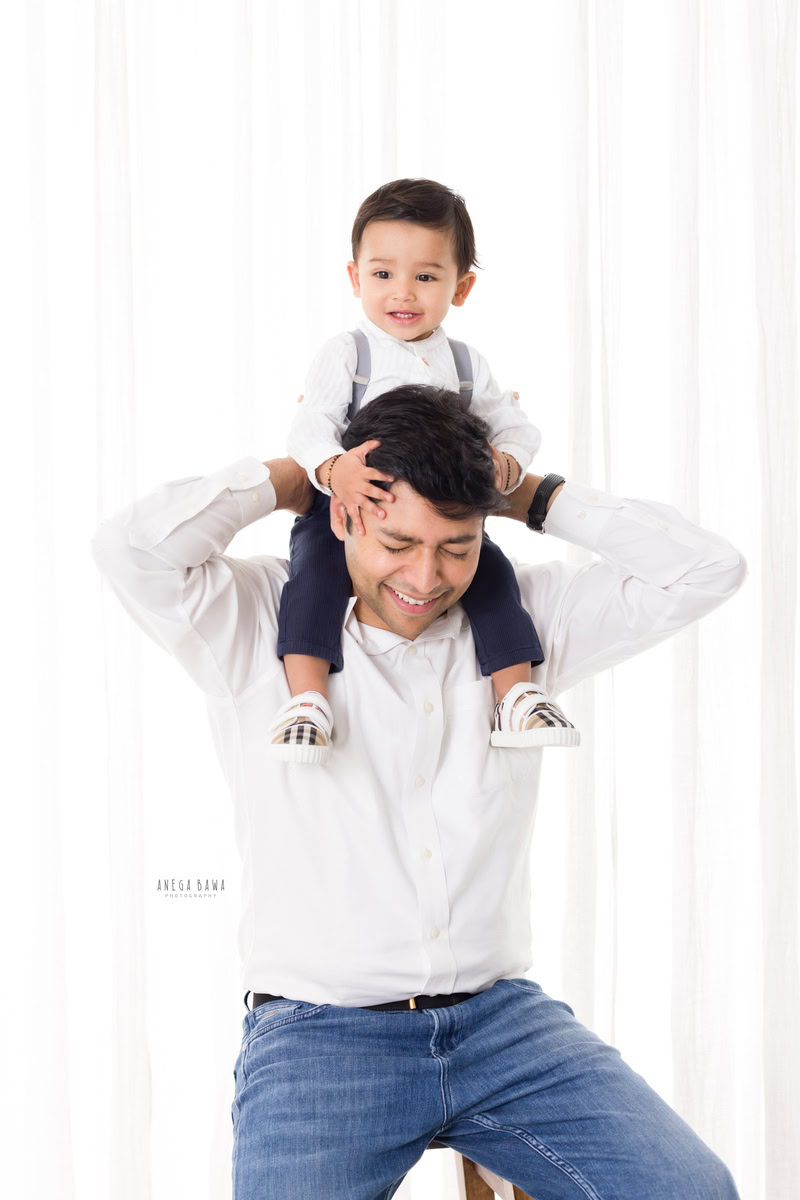 1-year-old boy seating on dad's shoulders against a white backdrop, both wearing white shirts, captured by Anega Bawa, family photographer in Gurgaon (Delhi NCR).