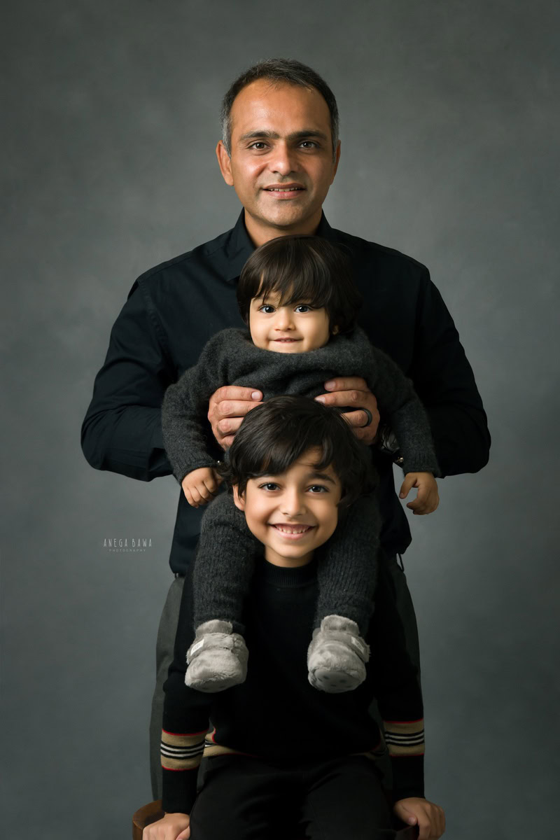 1-year-old boy seated on elder brother's shoulder with dad in the background, all wearing black against a grey backdrop, family photoshoot in Gurgaon (Delhi NCR)