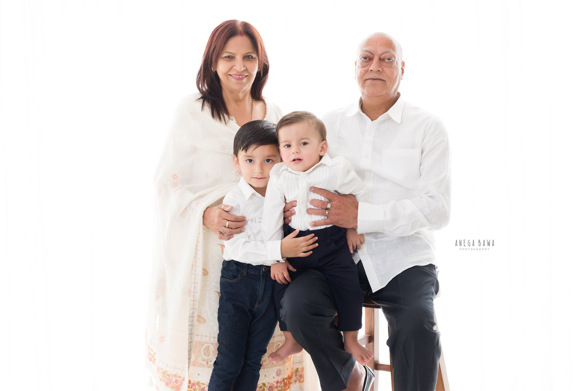 1-year-old boy seating on grandfather's lap with grandma and elder brother, against a white backdrop, captured by Anega Bawa, family photographer in Gurgaon (Delhi NCR).