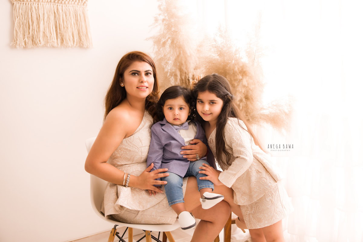 1-year boy seated on mom's lap with elder sister, set against a backdrop of bushes in beige, captured by Anega Bawa Photography for a family photoshoot in Gurgaon (Delhi NCR).