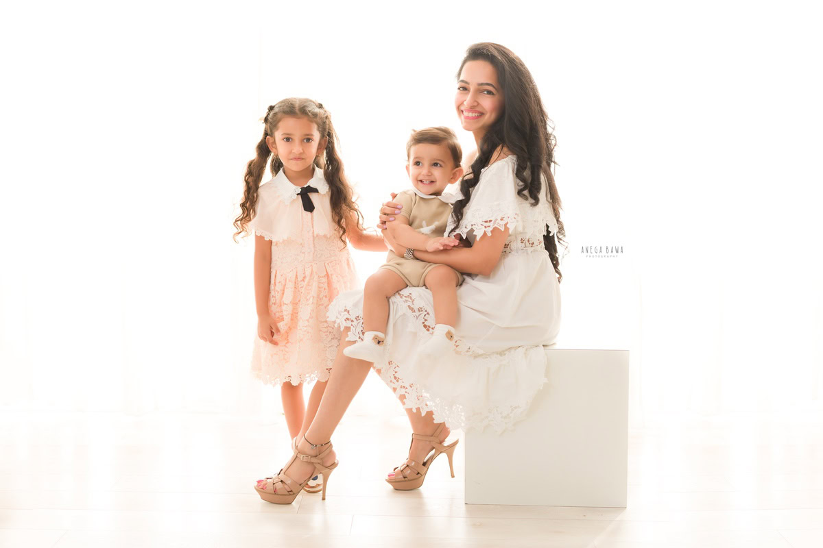 1-year-old boy seated on mom's lap, posing with his elder sister against a white backdrop, captured by Anega Bawa Family Photographer Gurgaon (Delhi NCR).