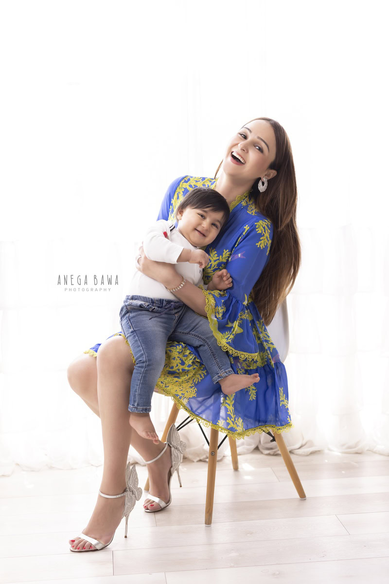 1-year-old boy seating on mom's lap in a smiling pose against a white backdrop, captured by Anega Bawa, family photographer in Gurgaon (Delhi NCR).