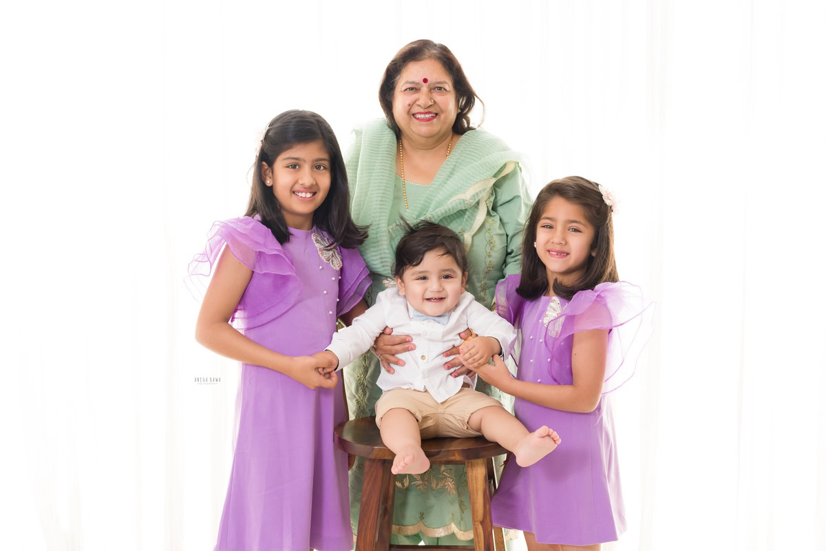 1-year-boy seated on a stool, posing with his sisters and granny against a serene white backdrop, captured by Anega Bawa Photography for a family photoshoot in Gurgaon (Delhi NCR).