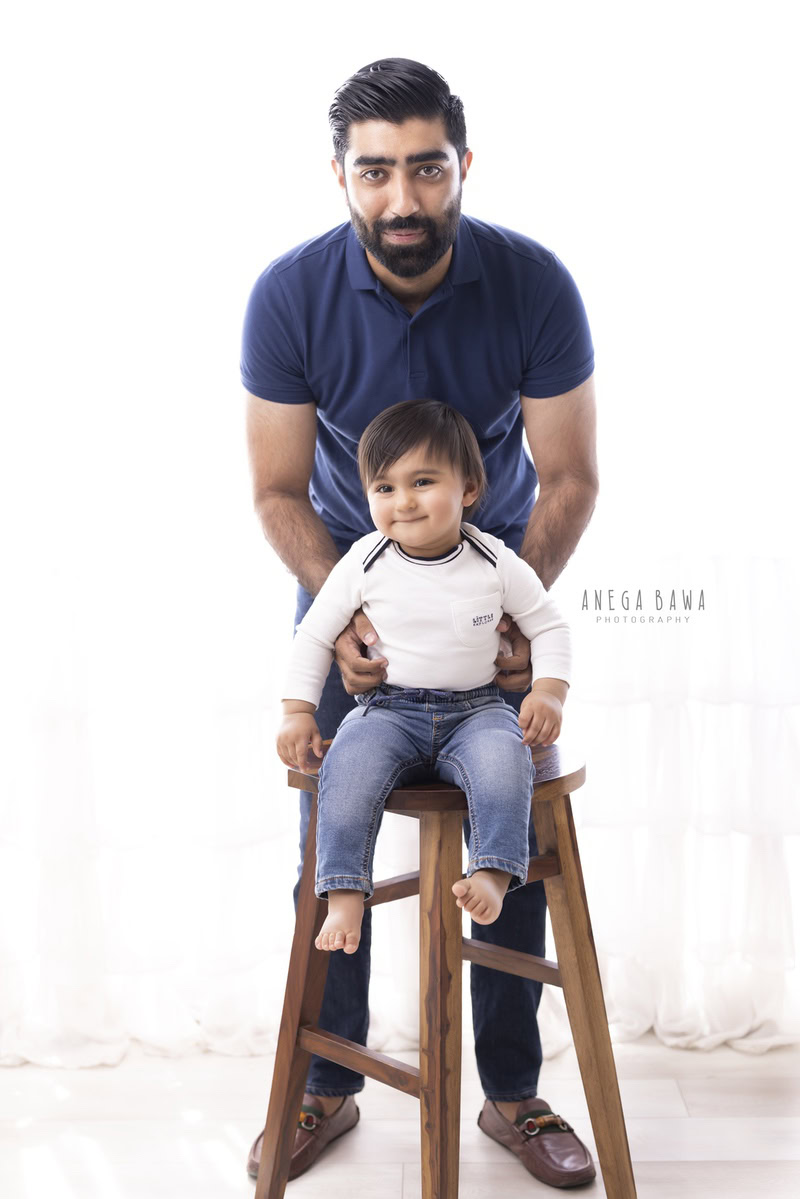 1-year-old boy seated on a wooden stool, posing with dad against a white backdrop, captured by Anega Bawa Photography for a family photoshoot in Gurgaon (Delhi NCR)