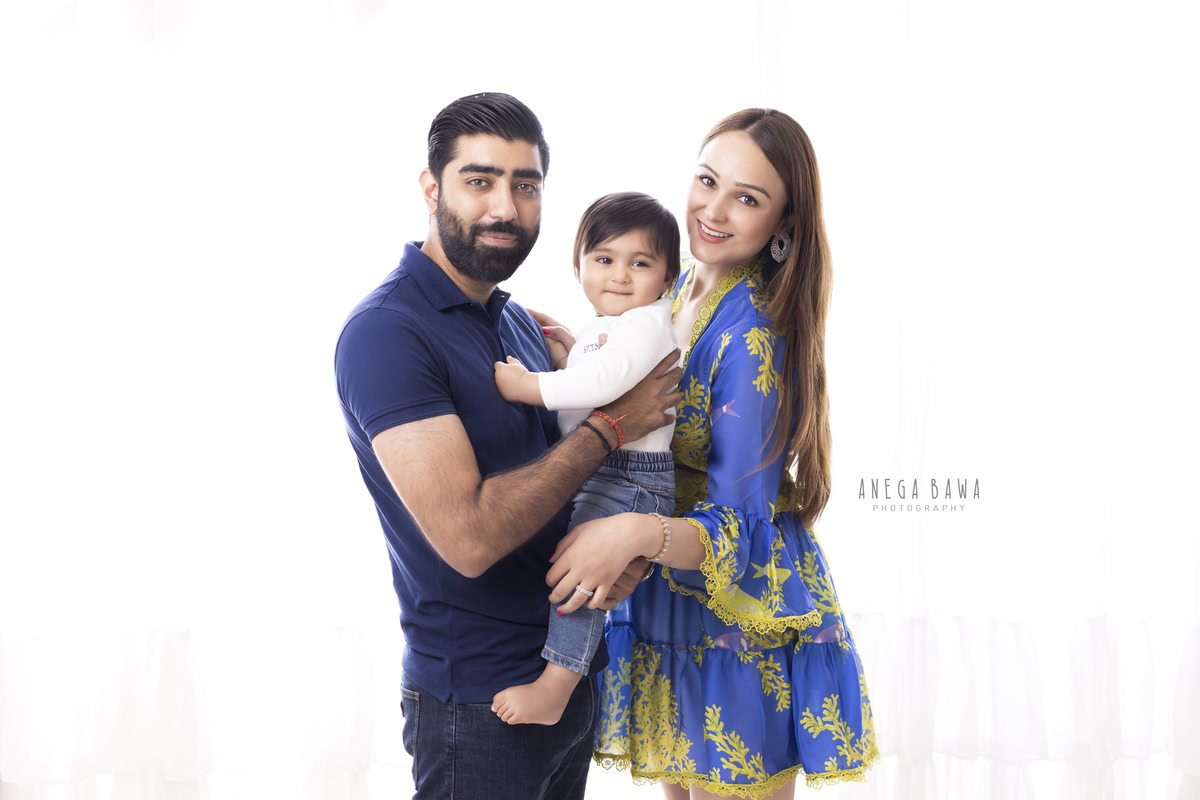 1-year-old boy smiling cutely with mom and dad against a white backdrop, captured by Anega Bawa Photography for a family photoshoot in Gurgaon (Delhi NCR).
