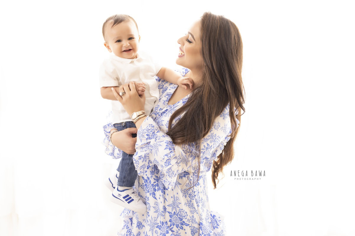 1-year-old boy smiling warmly in his mom's arms against a serene white backdrop, captured during a family photoshoot in Gurgaon (Delhi NCR).