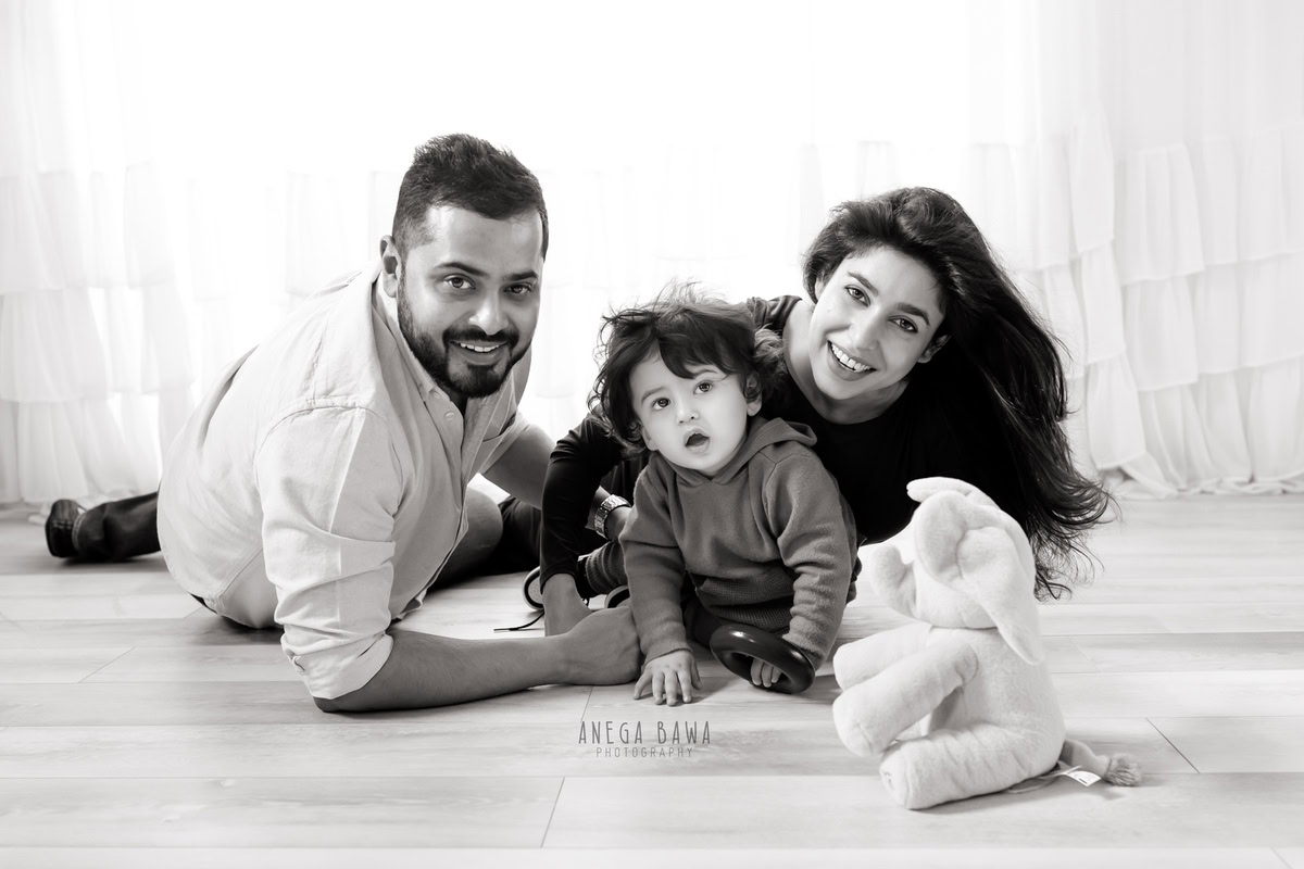 1-year-old boy smiling with mom and dad, lying on the floor with a soft toy against a white backdrop, captured by Anega Bawa Family Photographer Gurgaon (Delhi NCR).