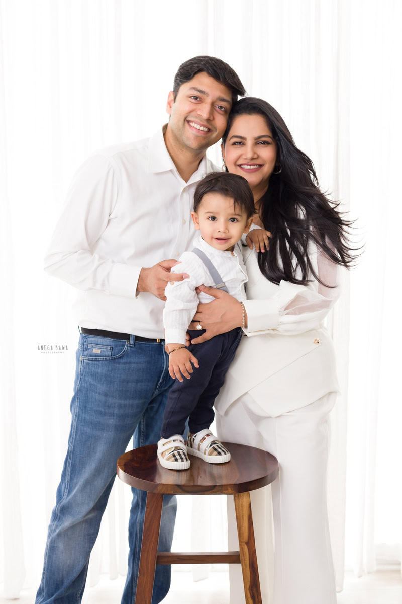 1-year-old boy standing cutely on a wooden stool, posing with his mom and dad against a crisp white backdrop, all dressed in white shirts.