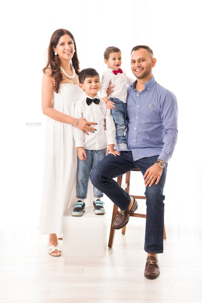 1-year-old boy standing on dad's lap, posing with elder brother and mom, against a white backdrop, family photoshoot in Gurgaon (Delhi NCR)